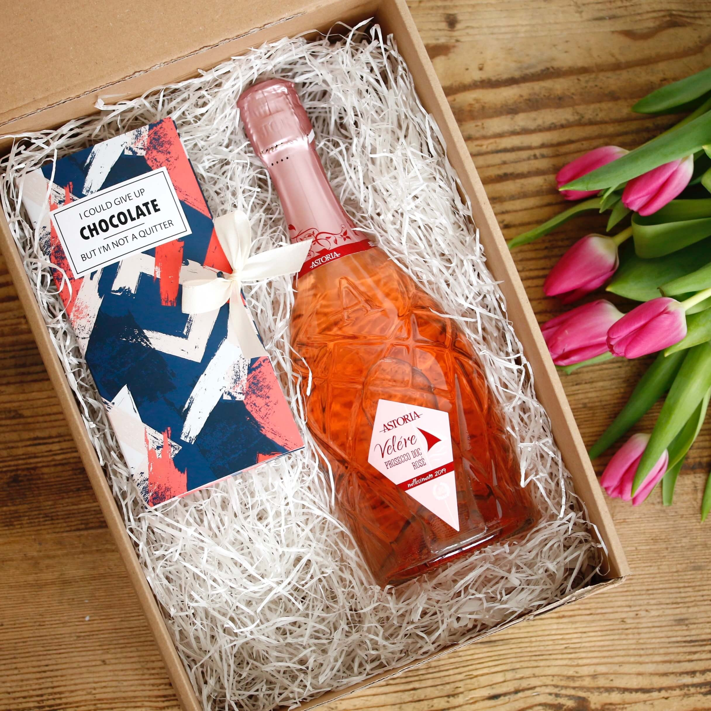 kartonska darilna škatla, v kateri je steklenica vina prosecco doc rose in čokolada v roza, belem in modrem zavitku s pentljo, na desni šopek roza tulipanov
